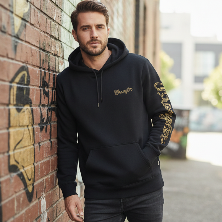 Image of Man wearing a black hoodie with gold text, leaning against a brick wall.