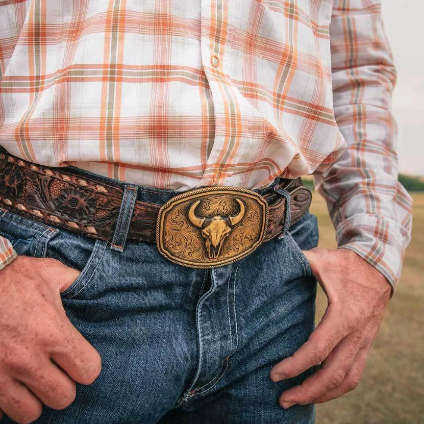 Image of Person wearing a plaid shirt, jeans, and a belt with a bull skull buckle in an outdoor setting.
