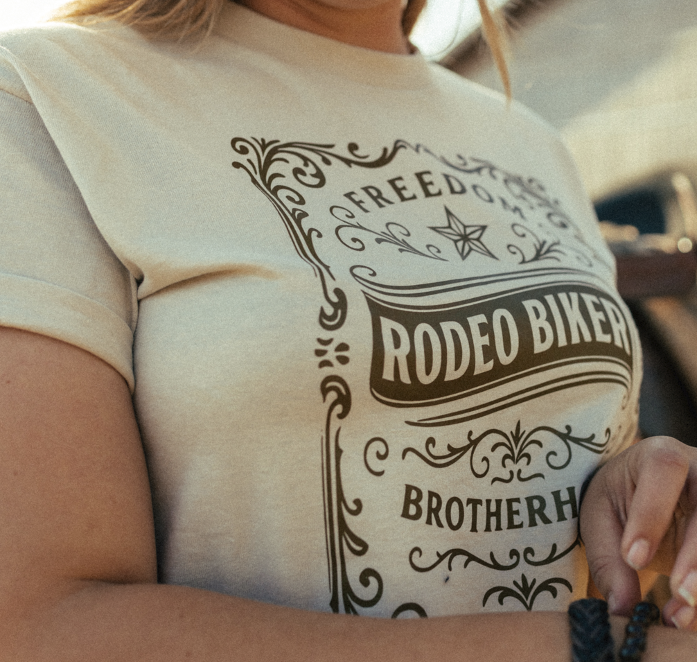 Image of A close-up of a person wearing a cream-colored t-shirt with a dark brown graphic that reads 'FREEDOM RODEO BIKER BROTHERHOOD' in an ornate vintage border design.