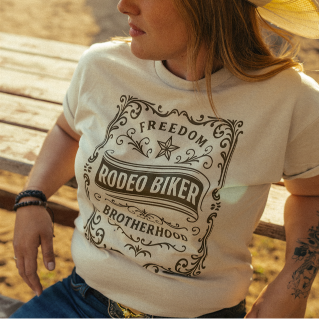 Image of A woman wearing a cream-colored graphic t-shirt with a western-themed 'Rodeo Biker Brotherhood' print, paired with a straw cowboy hat and blue jeans, sitting on a wooden bench outdoors.