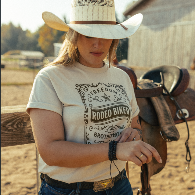 Image of A woman wearing a white cowboy hat and a cream graphic t-shirt that reads 'FREEDOM RODEO BIKER BROTHERHOOD' stands in a rustic outdoor setting next to a leather saddle.