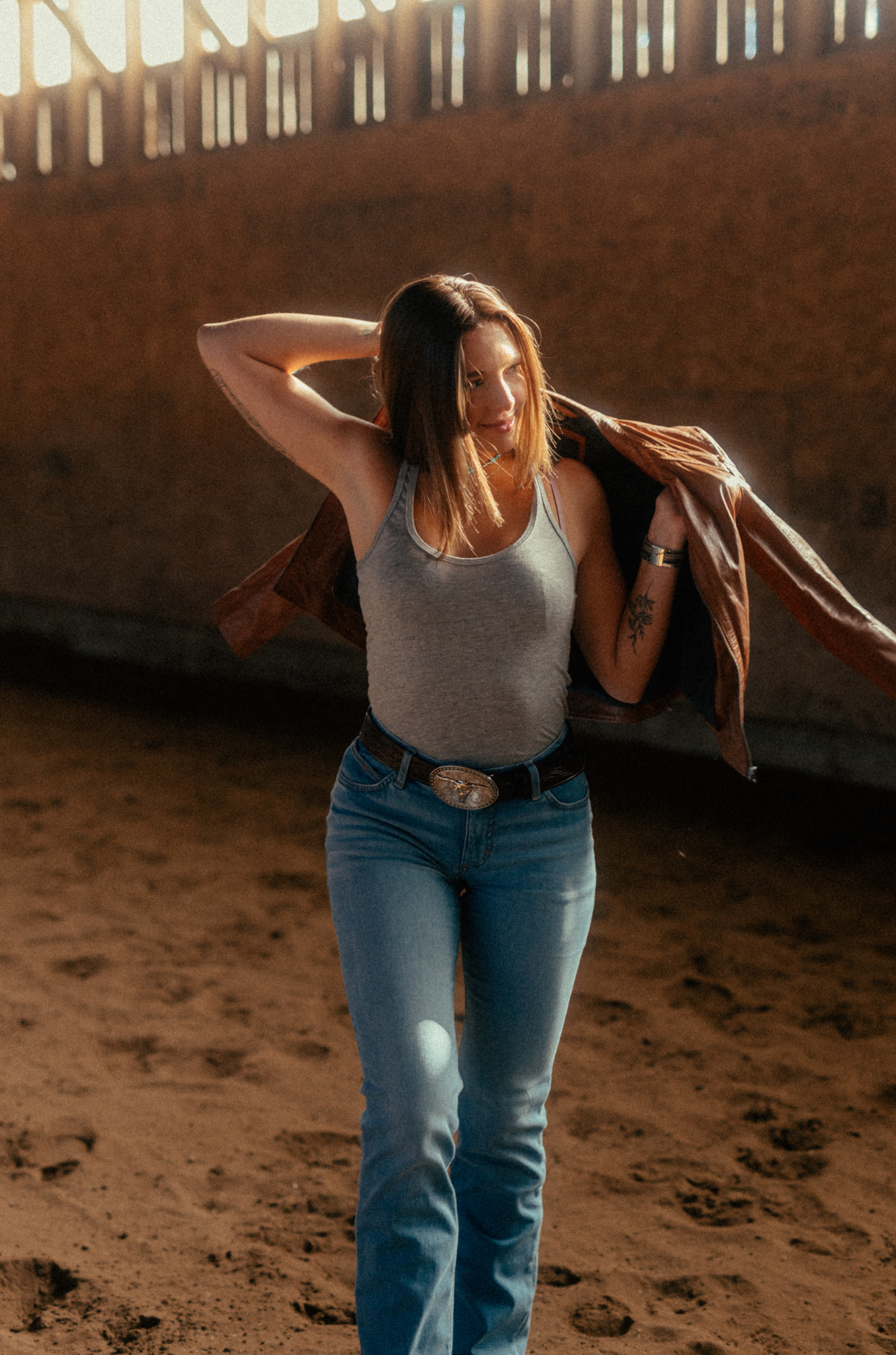 Woman in a tank top and jeans standing in a sunlit indoor arena.