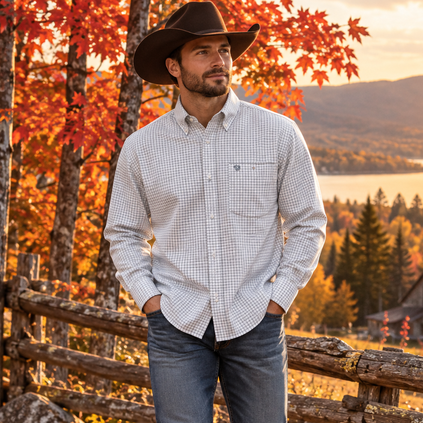 Image of A man wearing a light-colored gingham button-down shirt, blue jeans, and a black cowboy hat stands outdoors in an autumn setting with a wooden fence, colorful fall foliage, and a lake in the background.