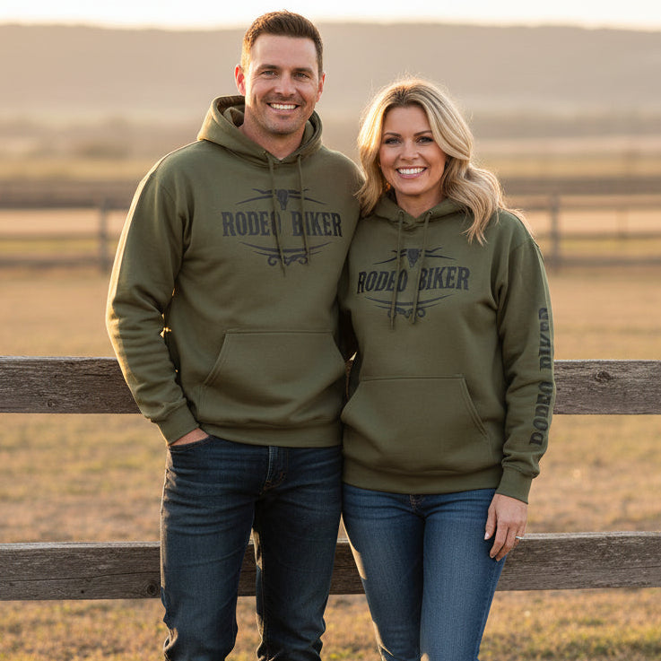 Image of Man and woman wearing matching green 'Rodeo Biker' hoodies standing in front of a wooden fence with a field in the background.