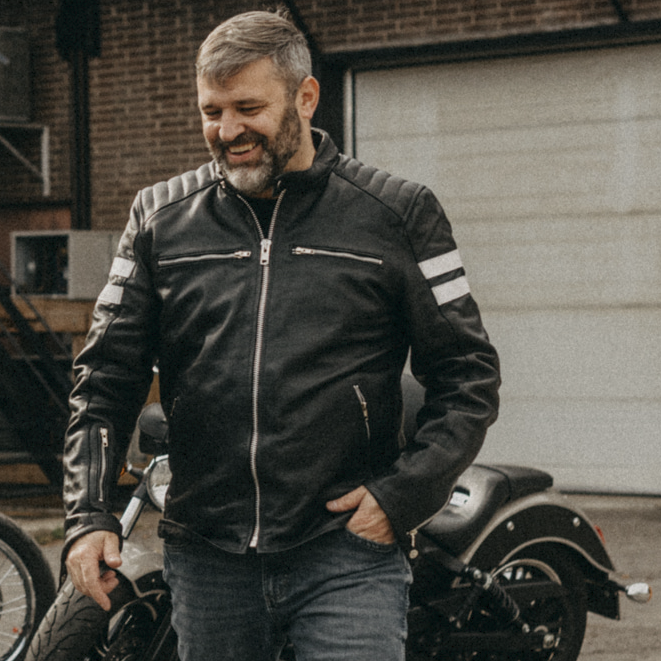 Image of Man wearing a black leather jacket with white stripes standing next to a motorcycle in an outdoor setting.