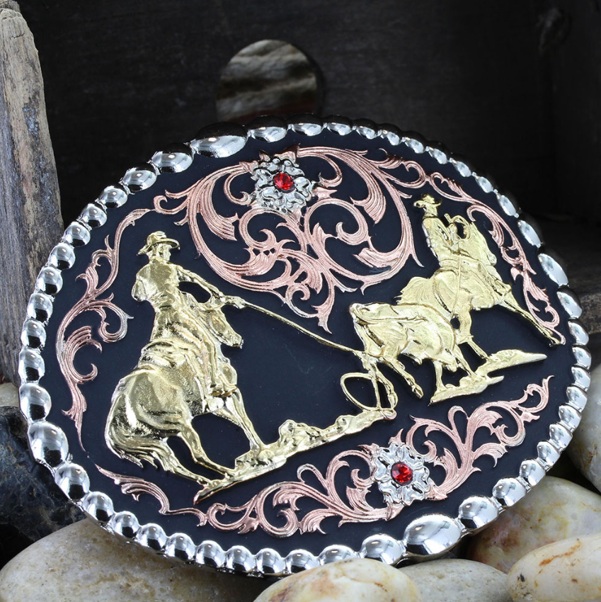 Image of An oval western-style belt buckle featuring a black enamel background with raised gold and rose gold filigree and figures on horseback, bordered by a scalloped silver-tone edge. Small red gem accents are visible near the top and bottom.