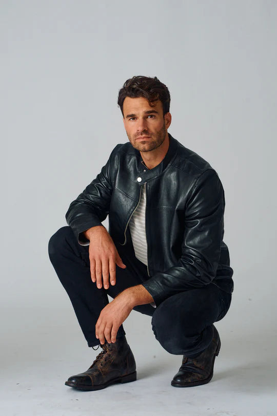 Image of Man wearing a black leather jacket and pants, crouching on a light gray background