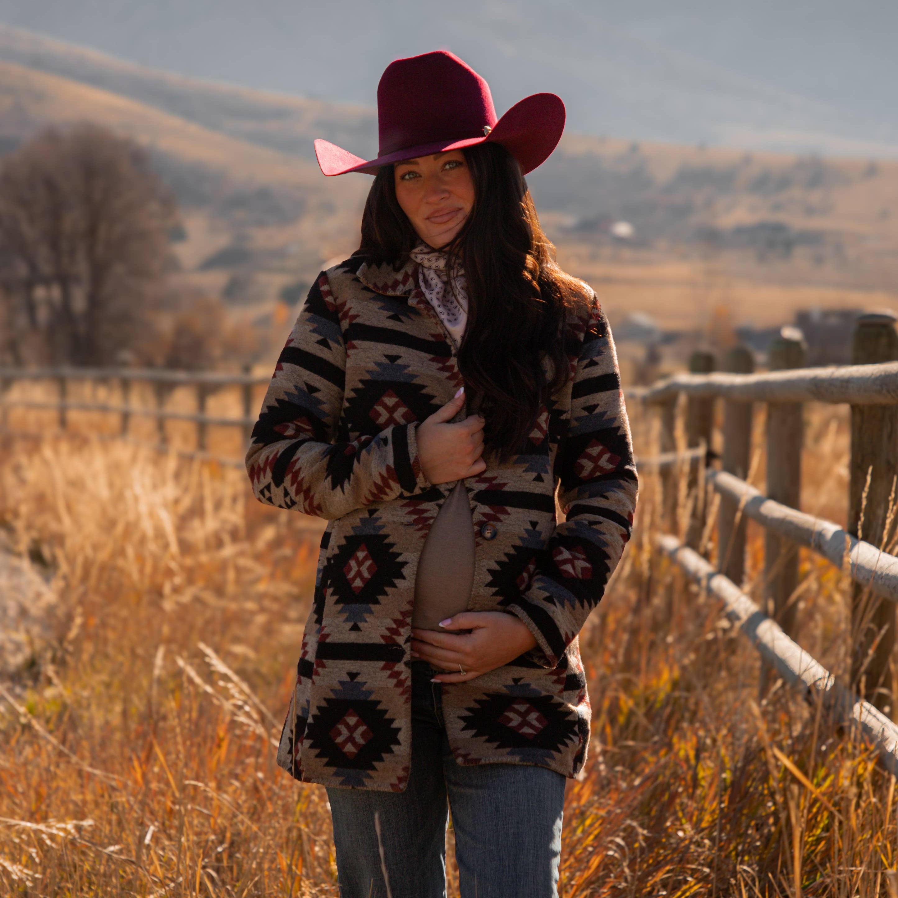 Image of A woman stands outdoors in a dry grassy field wearing a patterned Southwestern-style coat, blue jeans, a burgundy cowboy hat, and a patterned scarf, with mountains and a wooden fence in the background.