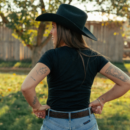 Image of Person wearing a black cowboy hat, black shirt, and blue jeans standing in a grassy field.