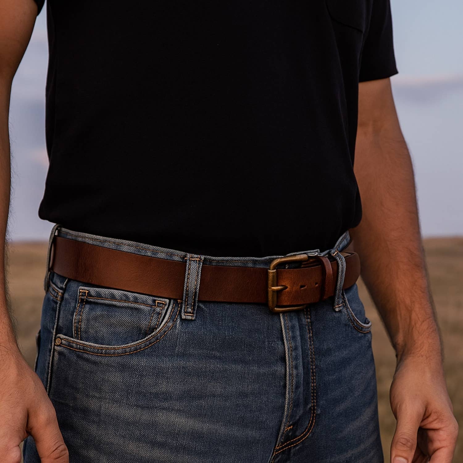 Image of Person wearing a black t-shirt, blue jeans, and a brown belt with a blurred natural background