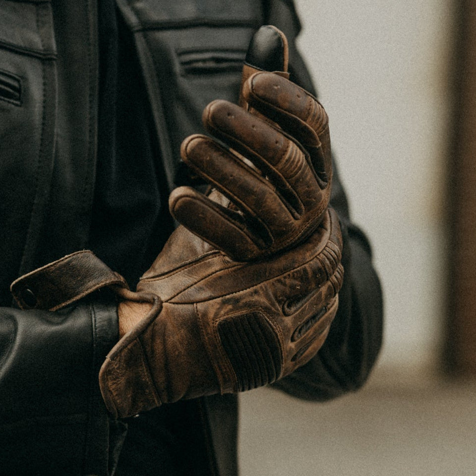 Image of Person wearing a black leather jacket and brown gloves, holding a motorcycle handle.