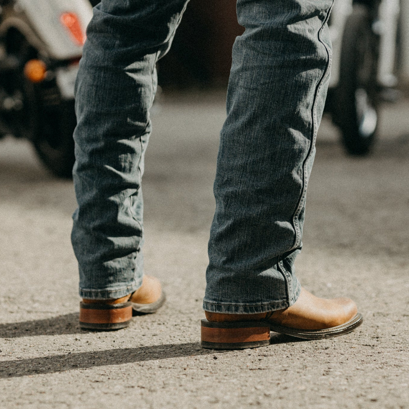 Image of Close-up of a person wearing blue jeans and brown boots on a street with motorcycles in the background.