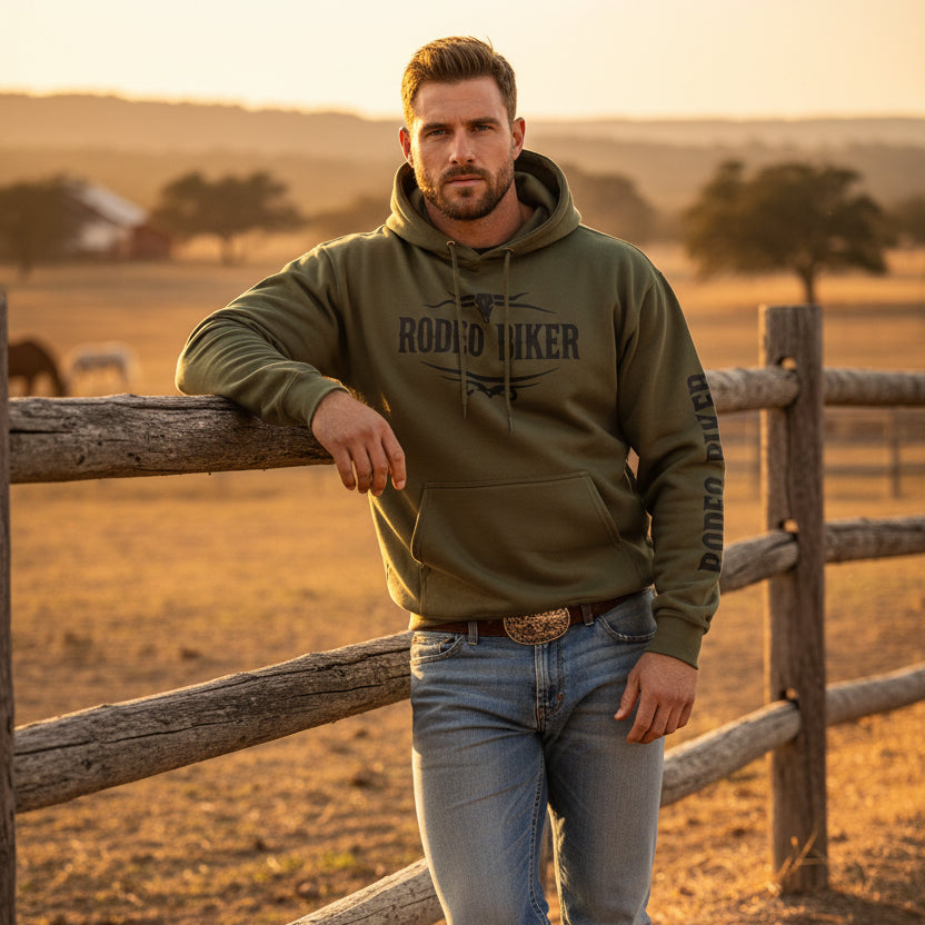 Image of Man wearing a green hoodie with 'Rodeo Biker' text, standing by a wooden fence in a rural setting.