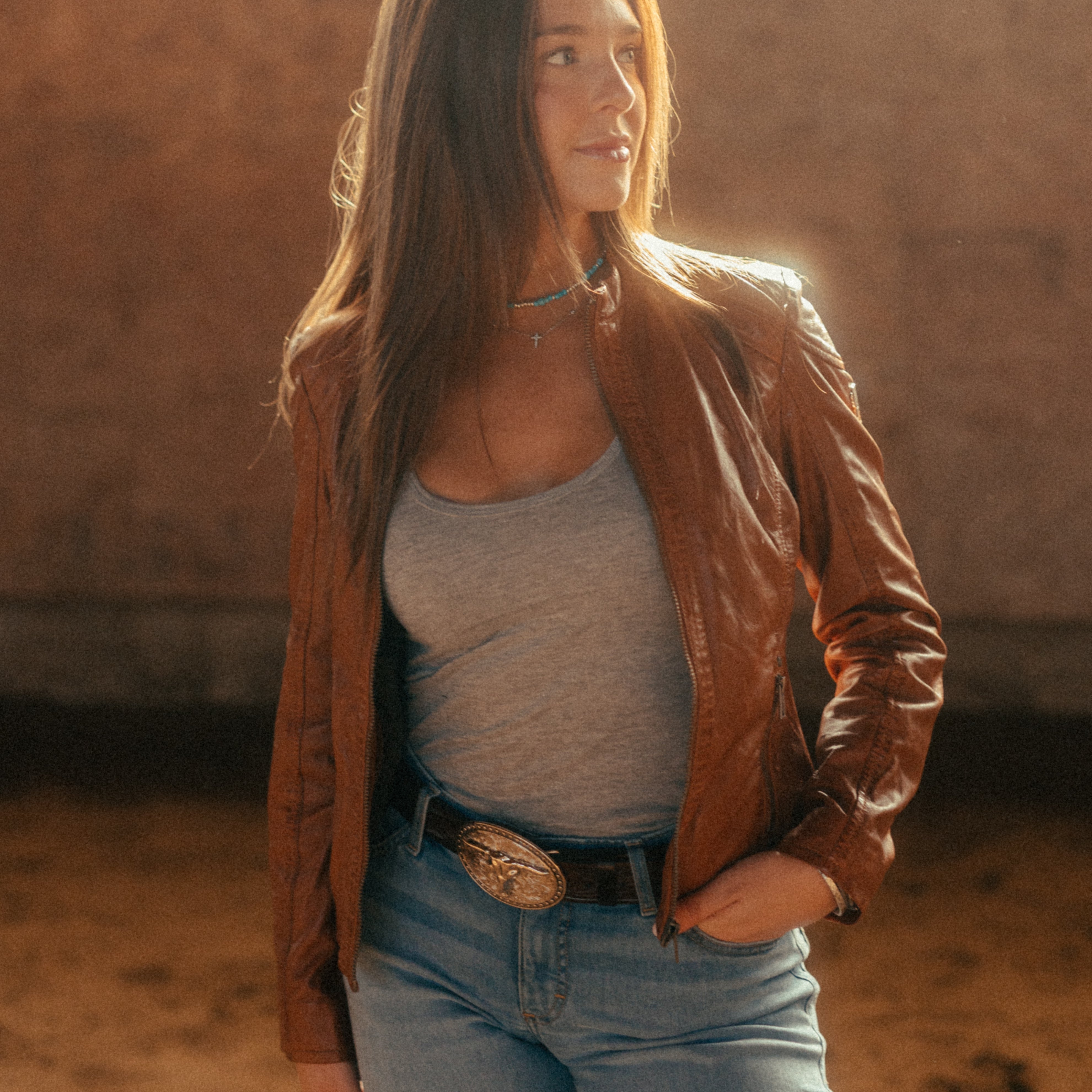 Image of Woman wearing a brown leather jacket and blue jeans standing in a desert-like setting with warm lighting.